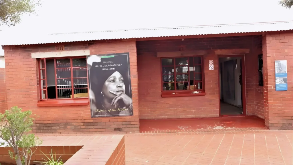Exterior of Winnie Madikizela-Mandela House in Soweto, Johannesburg, with a memorial portrait displayed on the brick facade.