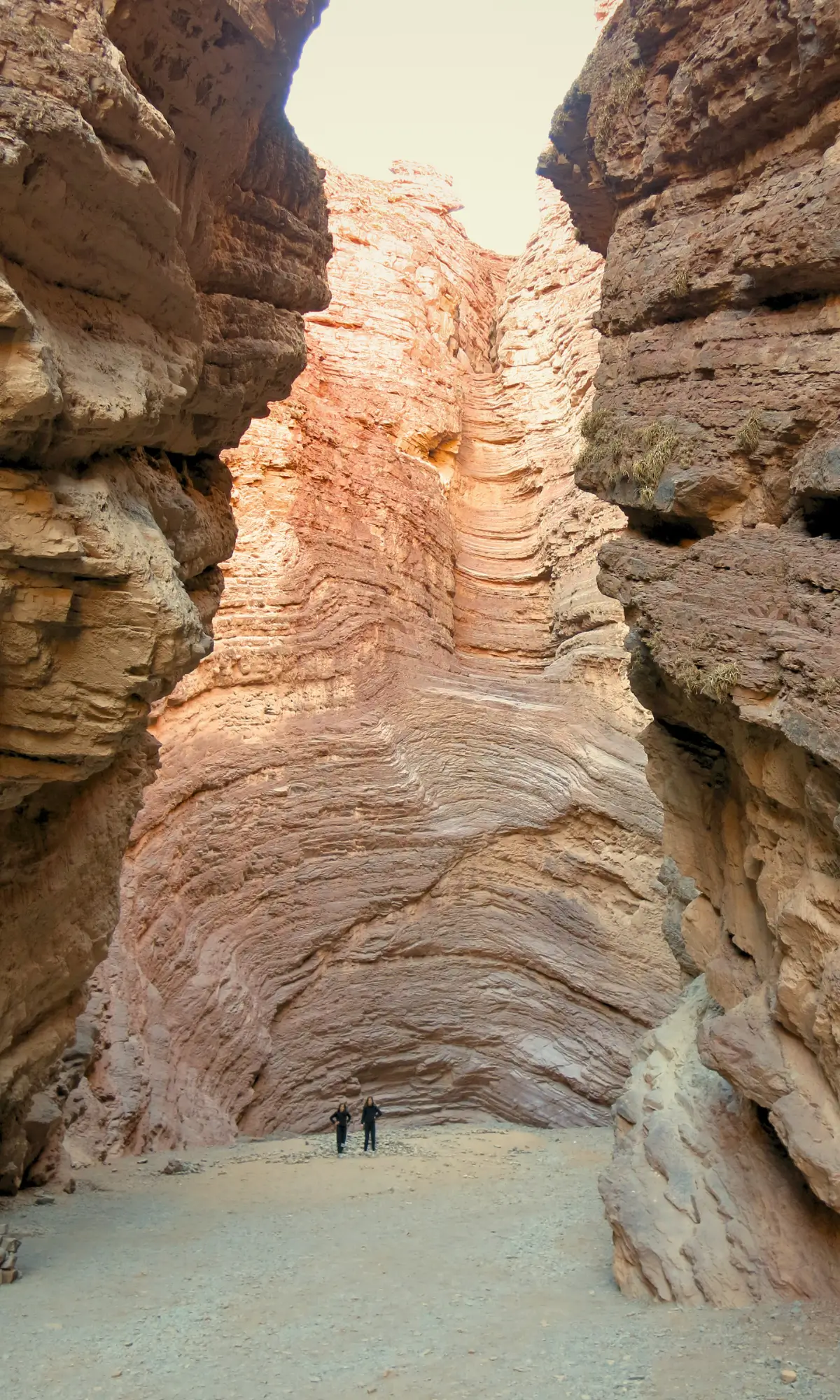 Two travellers standing inside a narrow canyon with layered red rock formations in Argentina.