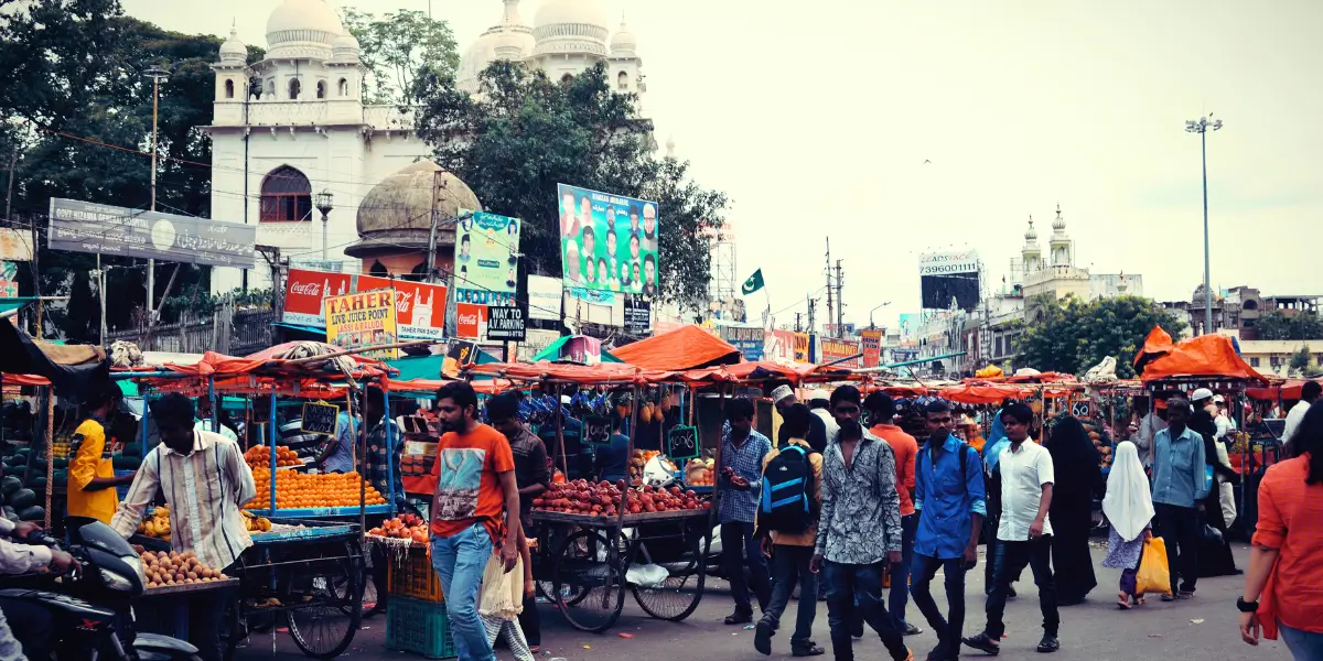 Busy street market in India with fruit vendors, pedestrians, and historic buildings in the background.