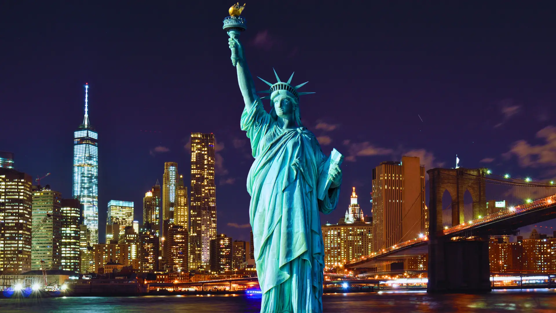 Statue of Liberty illuminated at night with the New York City skyline and Brooklyn Bridge in the background.