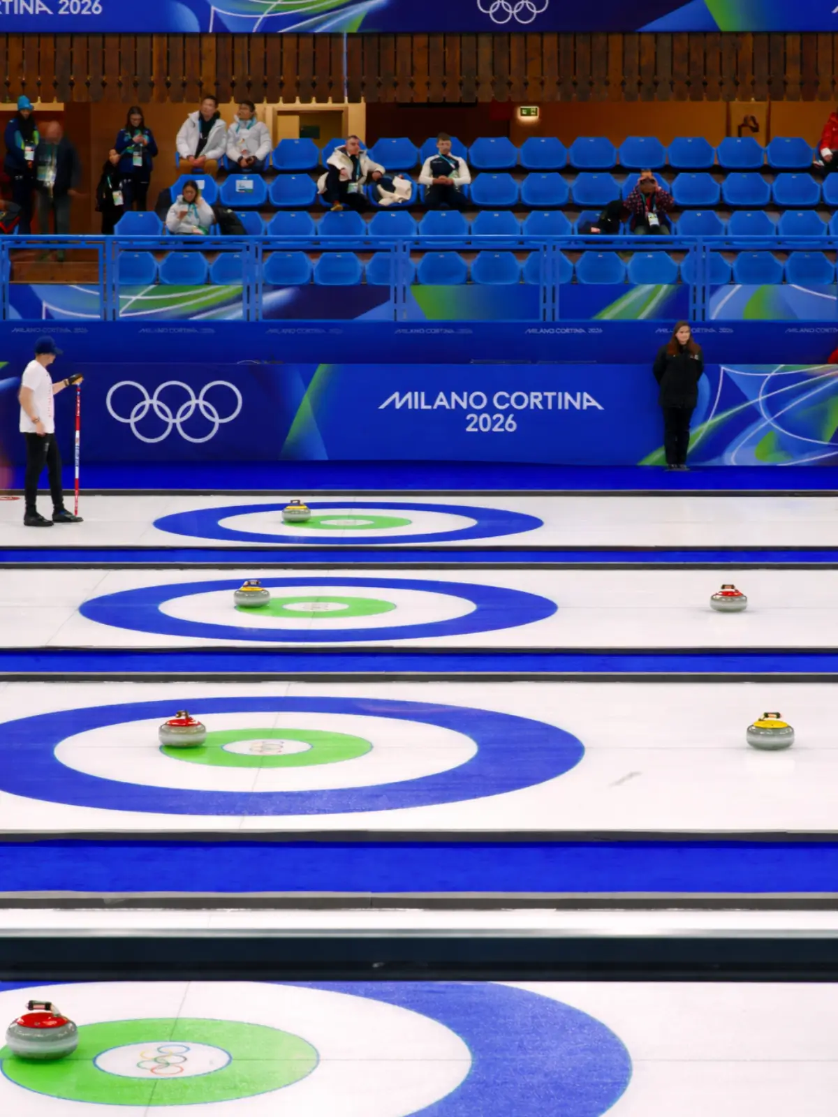 Curling match at Milano Cortina 2026 venue with Olympic rings and spectators in the stands.