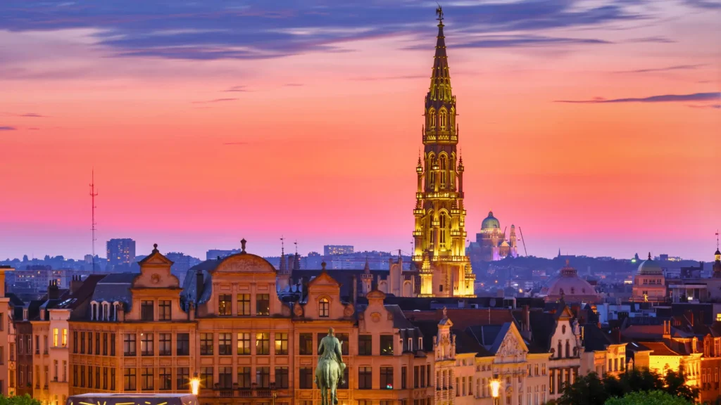 Brussels skyline at sunset with illuminated Gothic tower and historic rooftops in the city centre.