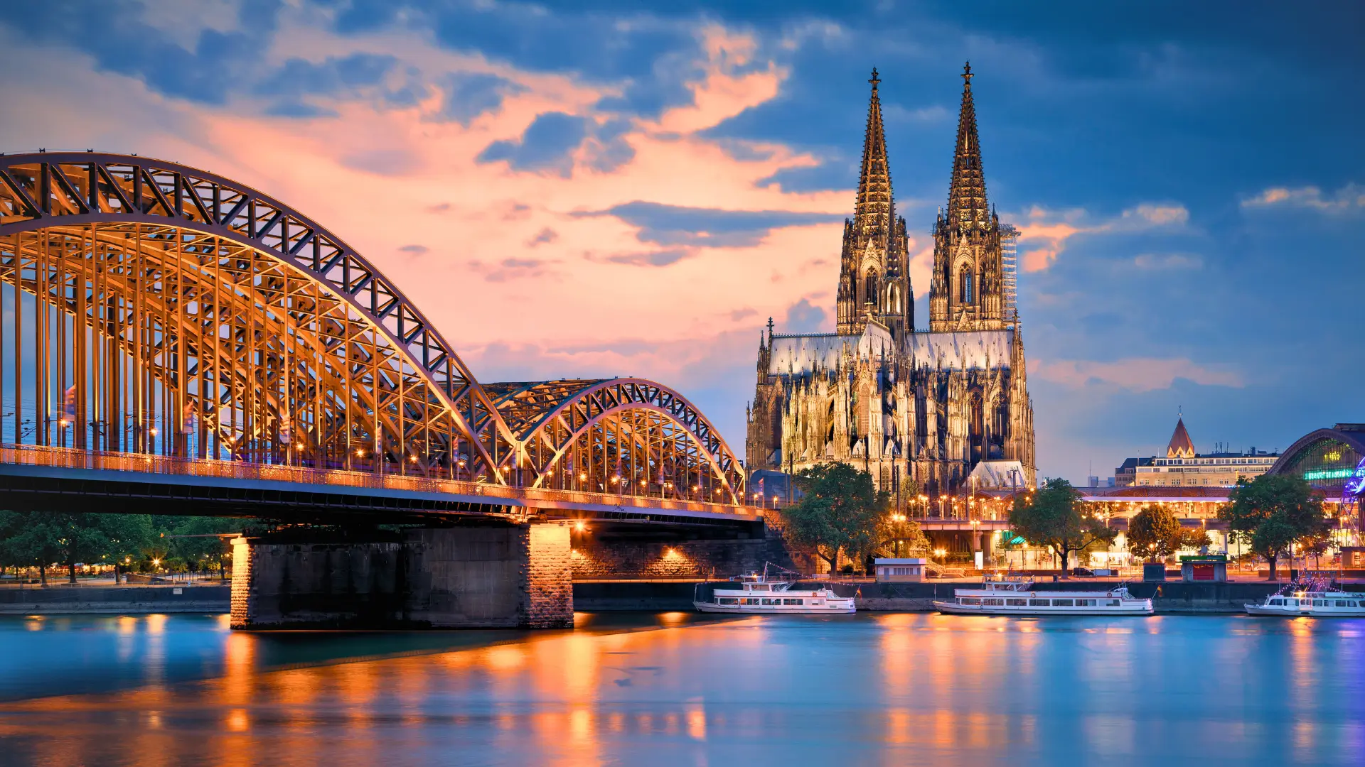 Cologne Cathedral and Hohenzollern Bridge over the Rhine River at sunset with city lights reflecting on the water.