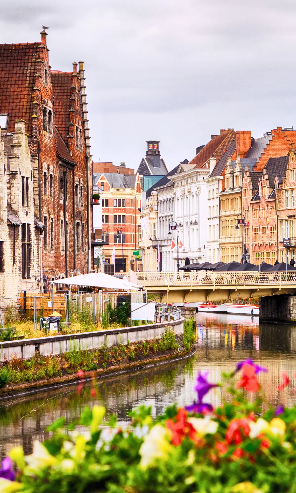 Historic buildings along a canal with a bridge and colourful flowers in Ghent, Belgium.