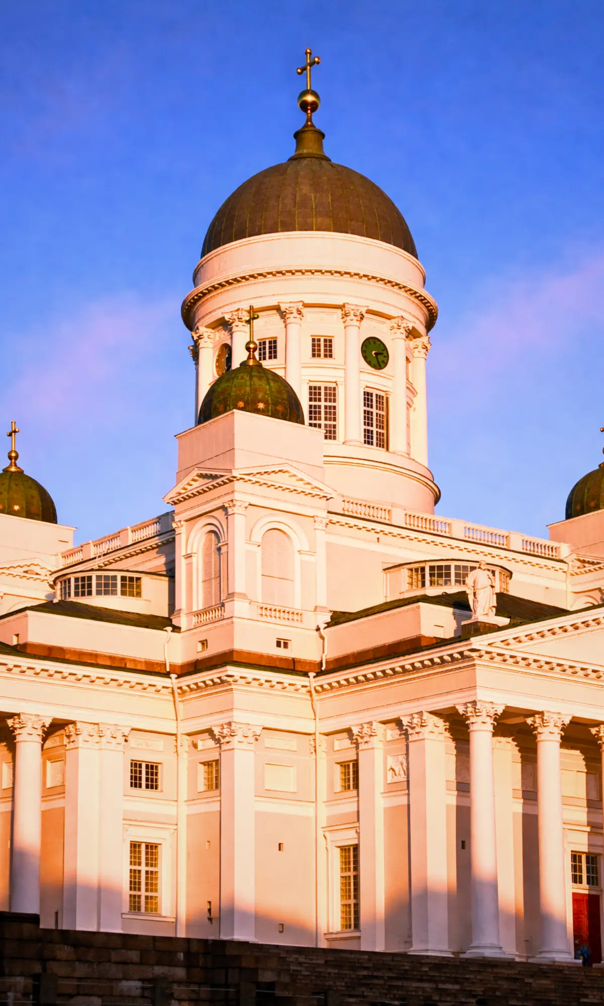 Helsinki Cathedral illuminated by warm sunset light with its white facade and green domes against a clear sky.