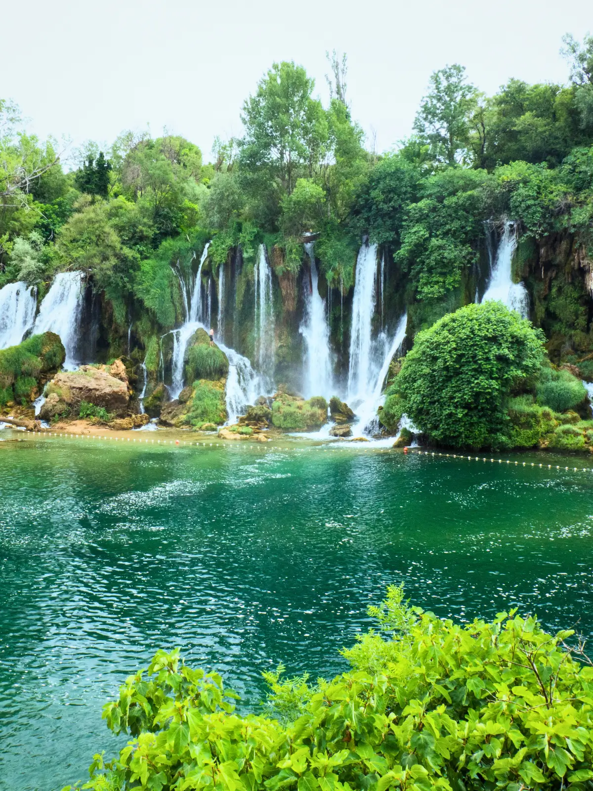 Kravica Waterfalls cascading into a turquoise pool surrounded by green forest in Bosnia and Herzegovina.