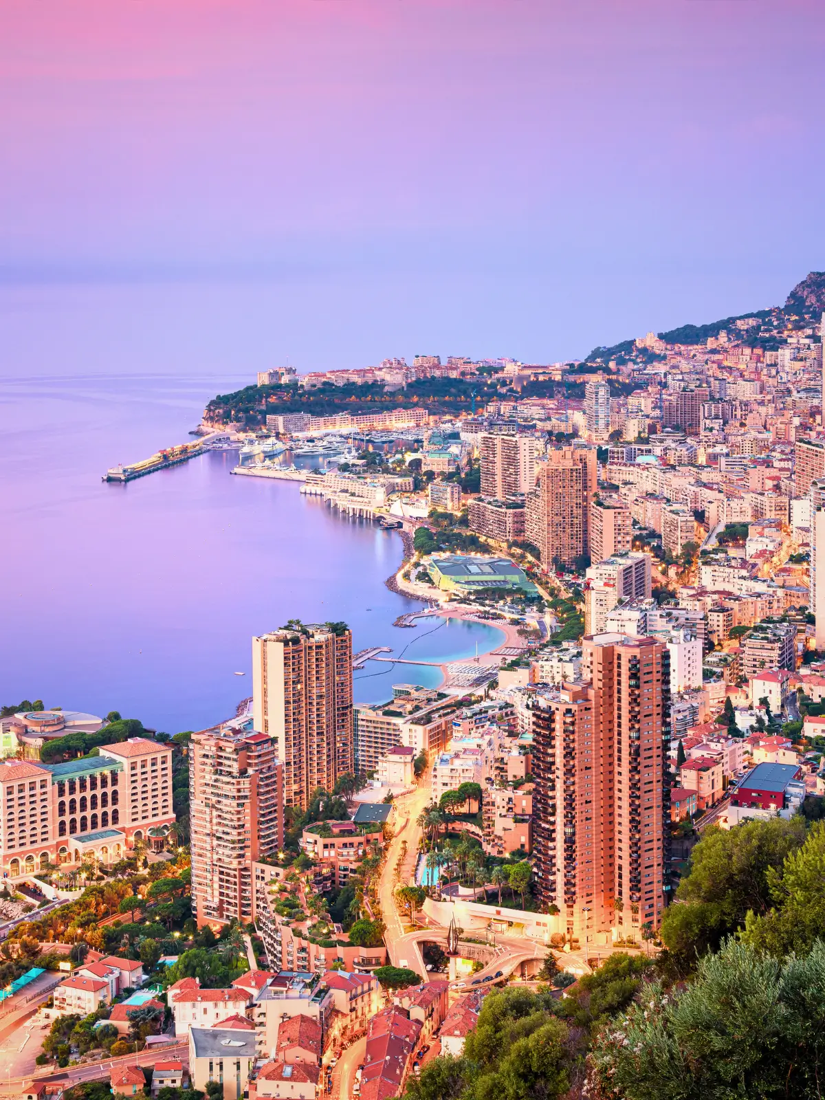 Aerial view of Monaco coastline at twilight with city lights, harbour, and high-rise buildings along the Mediterranean Sea.