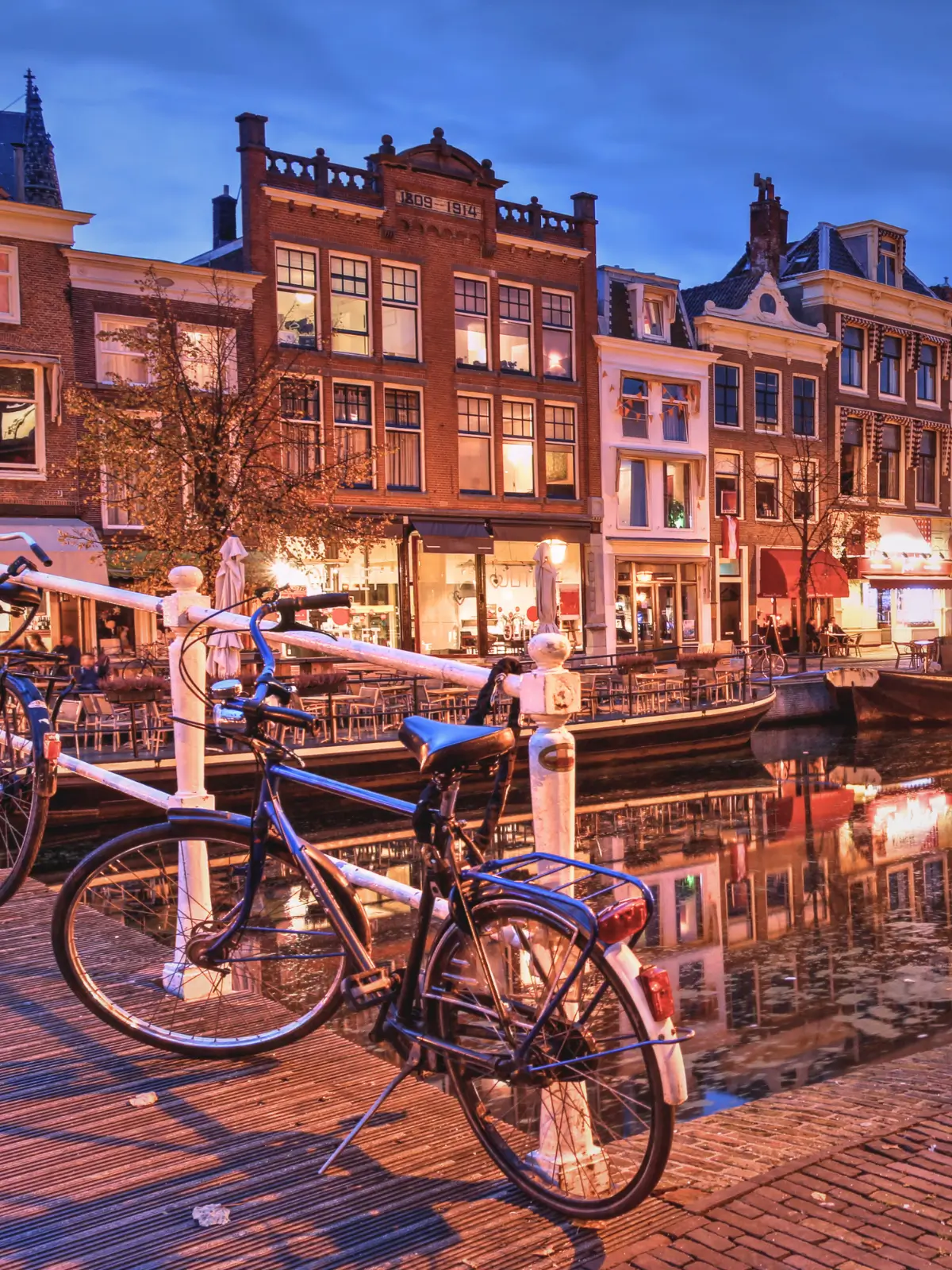 A bicycle parked beside a canal with illuminated buildings at dusk in Amsterdam.