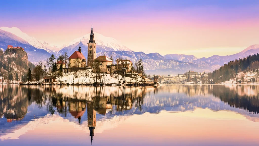 Lake Bled island with church and snow covered mountains reflected in calm water at sunset.