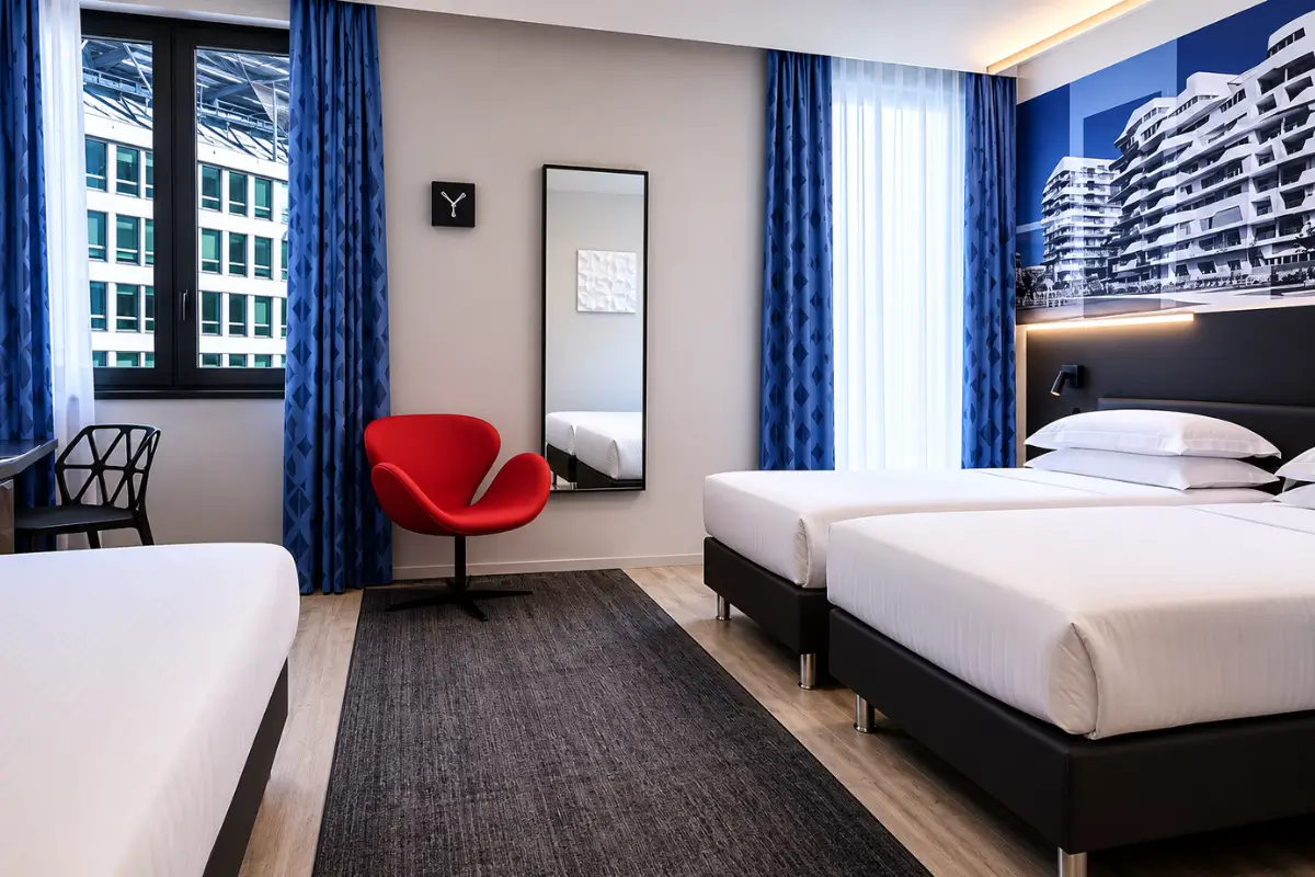 iQ Hotel Milano room with twin beds, blue curtains, and a red chair.