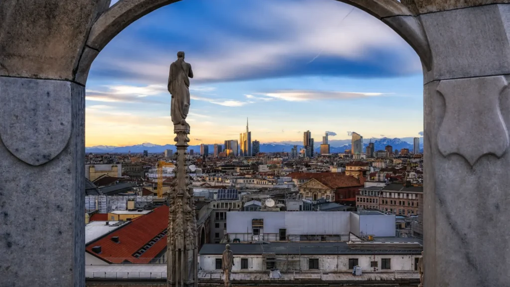 View of the Milan skyline framed by the arches and statues of the Duomo rooftop at sunset.