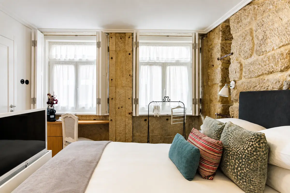 Bright bedroom at 1872 River House by Olivia with exposed stone walls, white shutters, and a neatly made bed.