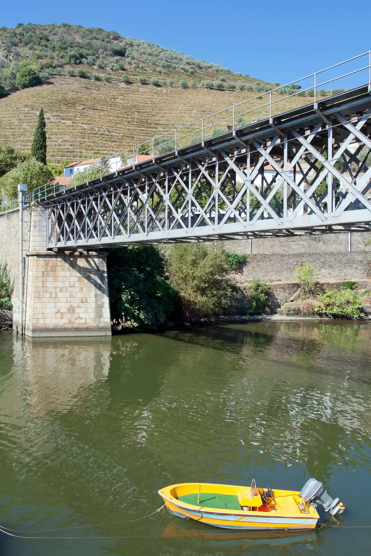 Metal bridge over the Pinhão River with a small boat and terraced vineyards behind it.
