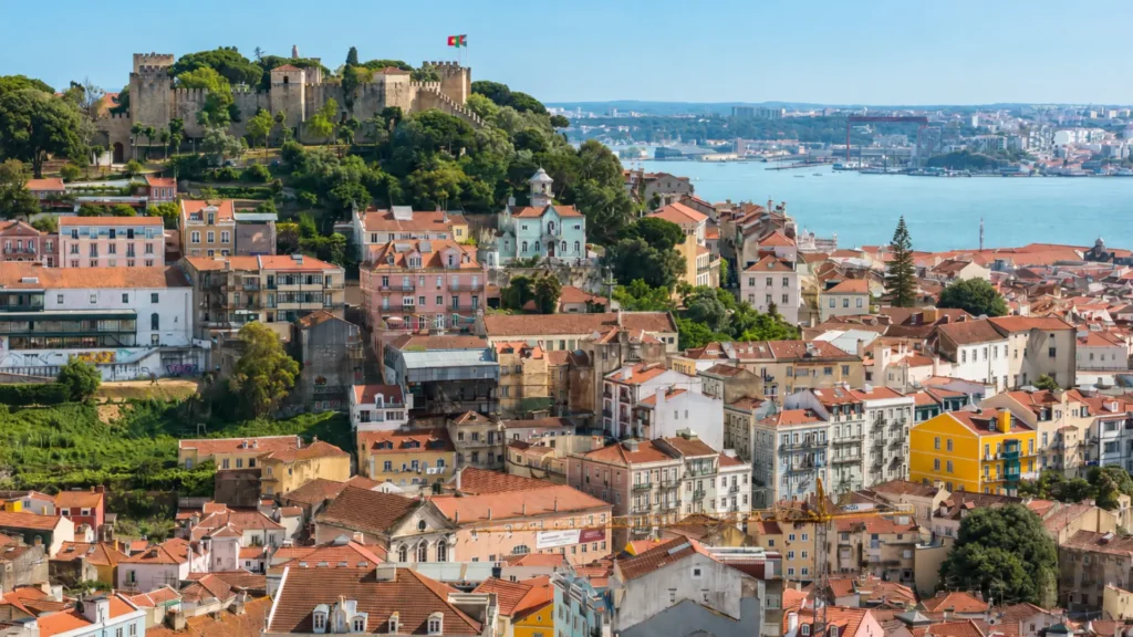 Panoramic view of Castelo de São Jorge on a green hilltop overlooking traditional terracotta rooftops and the Tagus River in Lisbon.