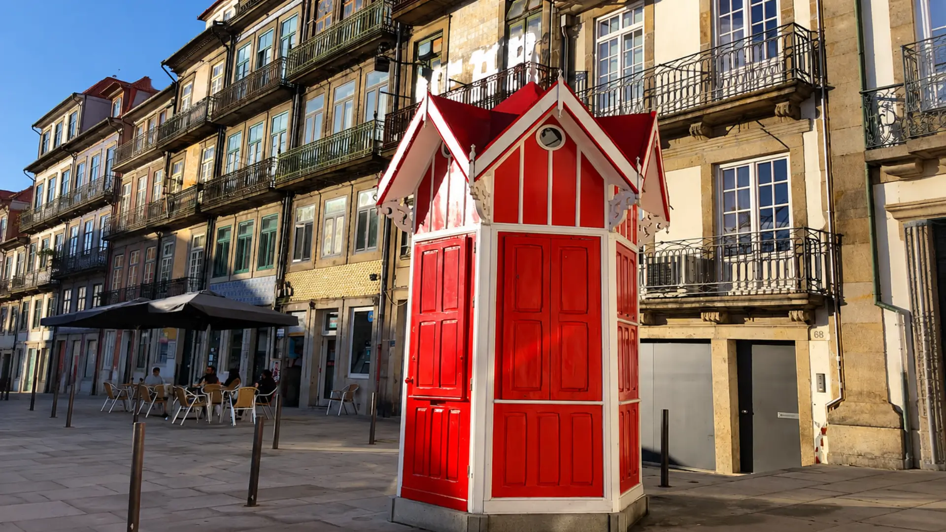 Red and white kiosk in Cedofeita, Porto, surrounded by historic buildings with wrought-iron balconies.