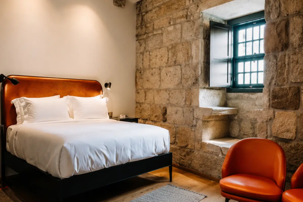 Bedroom at Exmo. Hotel in Porto with an exposed stone wall, white bed and orange chairs.