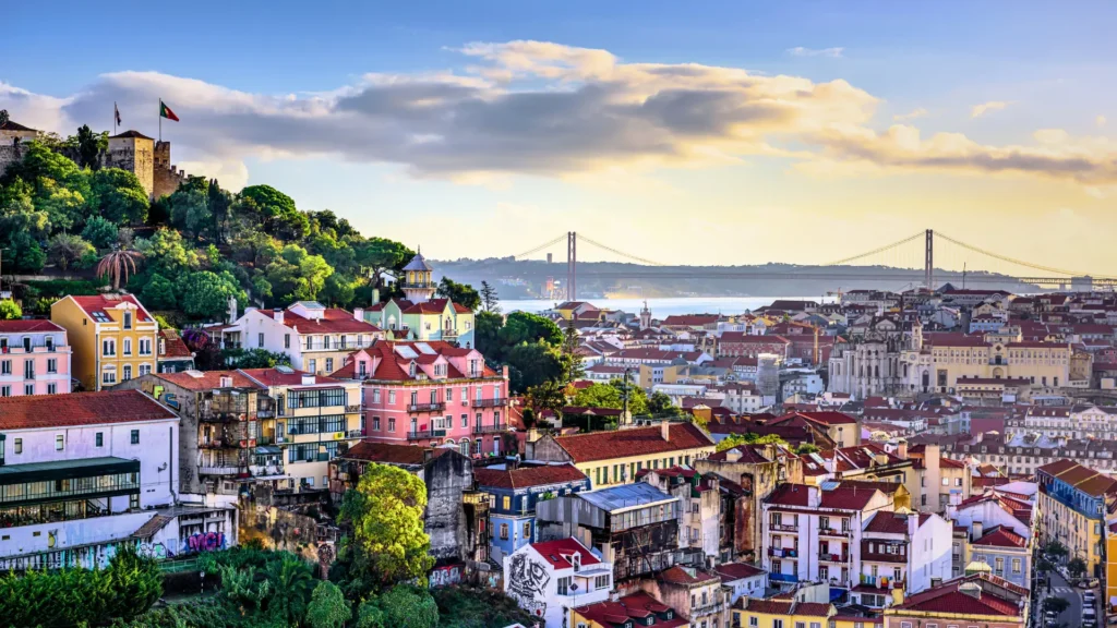 Panoramic view of Lisbon featuring colourful terracotta-roofed buildings, São Jorge Castle on the hilltop, and the 25 de Abril Bridge in the distance under a warm sky.