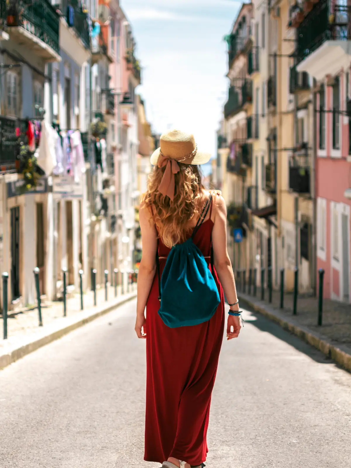 A female tourist with a straw hat and backpack walking down a narrow, cobblestone street in Lisbon.