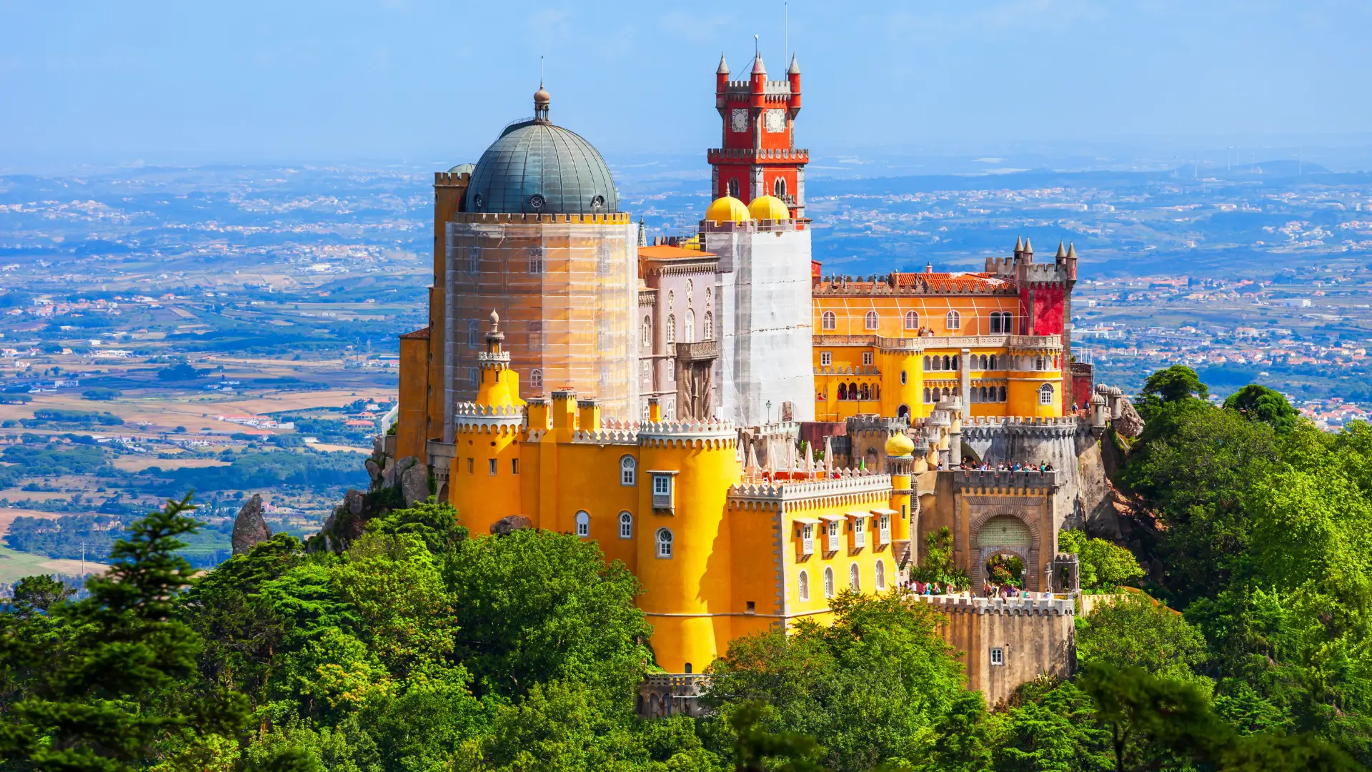 Panoramic view of the colourful yellow and red Pena Palace sitting atop a lush green mountain in Sintra under a clear blue sky.