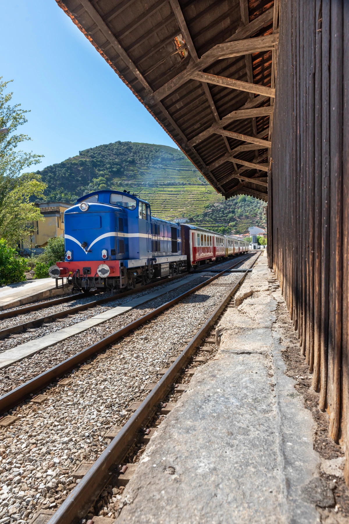Train beside the platform at Pinhão Railway Station with vineyard-covered hills behind it.